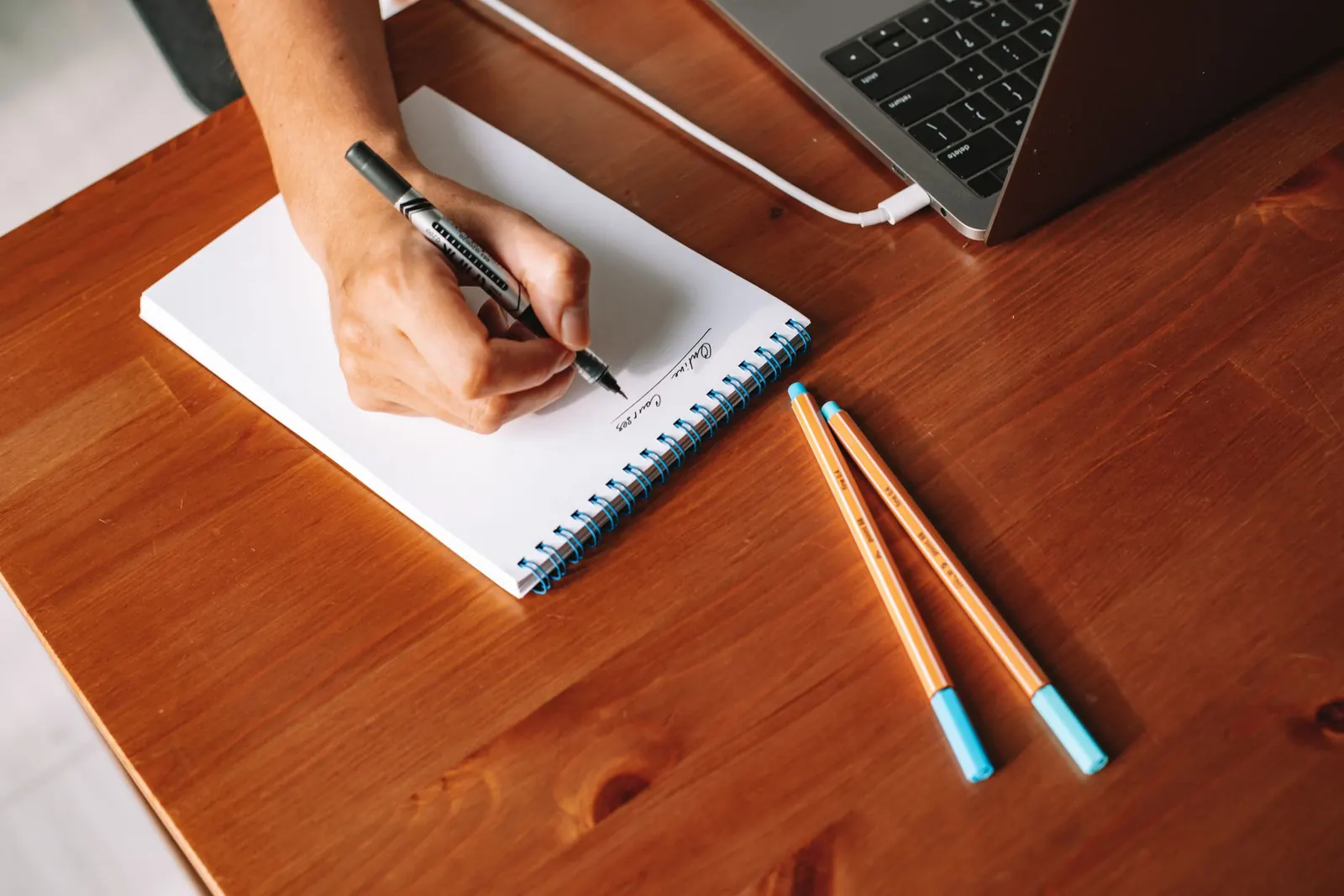 A hand writing in a paper notebook on a wooden desk in afternoon light